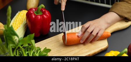 Vue rapprochée de la cuisinière à couper des carottes fraîches sur le bloc à hacher dans une cuisine moderne sombre Banque D'Images