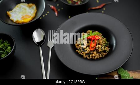 Cuisine traditionnelle thaïlandaise, porc haché frits au basilic (Pad ka prao), œufs frits et sauce au Chili servis sur des assiettes en céramique noire dans le restaurant thaïlandais Banque D'Images