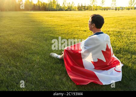Concept de la fête du Canada. Les hommes d'âge moyen sont assis sur l'herbe dans le parc avec le drapeau canadien sur son dos. Concept de vacances nationales Banque D'Images