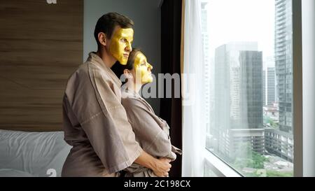 un jeune homme en peignoir câlins petite amie avec masque facial doré près d'une grande fenêtre avec des gratte-ciel dehors dans une chambre d'hôtel moderne Banque D'Images