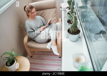 Une fille blonde lit le livre tout en étant assise sur un fauteuil beige appartement Banque D'Images