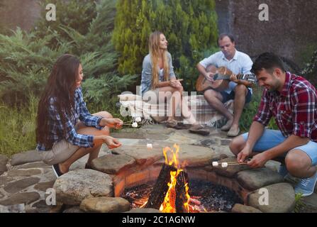 Compagnie de jeunes tenant des bâtons avec des guimauves au-dessus du feu de camp dans la soirée Banque D'Images