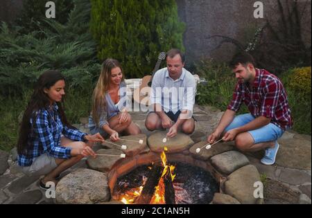 Compagnie de jeunes tenant des bâtons avec des guimauves au-dessus du feu de camp dans la soirée Banque D'Images