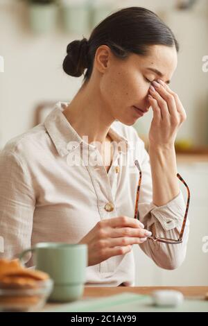 Portrait vertical à tons chauds de la jeune femme fatiguée qui se frotte contre le pont du nez et qui prend des lunettes tout en souffrant de maux de tête Banque D'Images