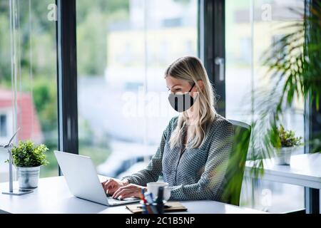 Jeune femme avec masque de visage au travail après avoir été verrouillée, en utilisant un ordinateur portable. Banque D'Images