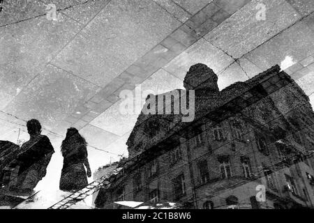 Silhouettes ombrées et réfléchies dans une flaque d'un bâtiment néoclassique et deux personnes marchant sur une rue piétonne humide, en noir et blanc Banque D'Images