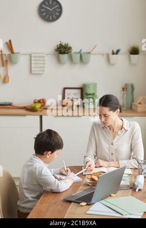 Portrait vertical aux tons chauds de la jeune mère et du fils qui font leurs devoirs tout en étant assis à une table dans une cuisine confortable, un espace pour les copies Banque D'Images