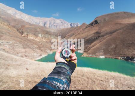 La main d'un homme tient un compas magnétique de poche pour la navigation sur fond de pente rocheuse et d'un lac de montagne. La conce Banque D'Images