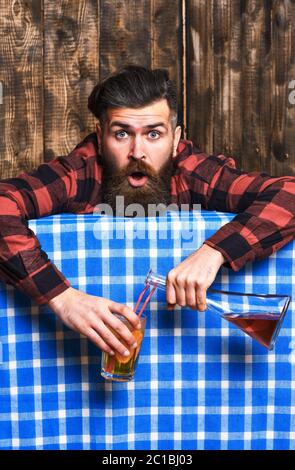 Homme en chemise à carreaux sur fond de bois, nappe bleue. Concept de gueule de bois. Barman avec barbe et visage choqué tient le verre de cocktail et la bouteille avec l'alcool dans les mains. Boisson alcoolisée Barman, en état d'ivresse. Banque D'Images