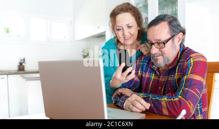Un couple âgé utilise simultanément son smartphone et son ordinateur portable depuis sa cuisine à la maison. Restez à la maison et nouveau concept de communication. Banque D'Images
