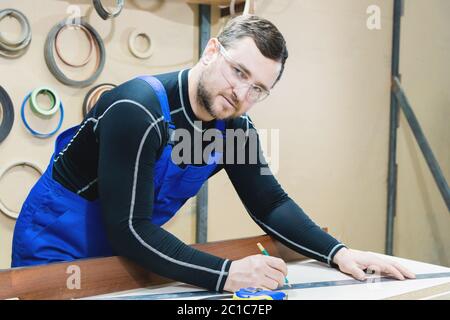 Un charpentier barbu fatigué sur une table avec un crayon dessine une enseigne sur le tableau. Sur le lieu de travail il y a un mas ordinaire Banque D'Images