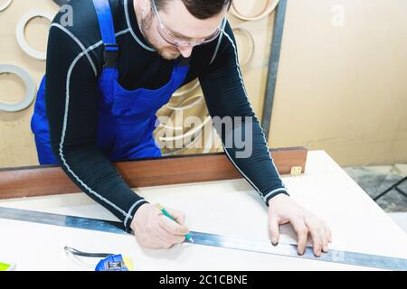 Un charpentier barbu fatigué sur une table avec un crayon dessine une enseigne sur le tableau. Sur le lieu de travail il y a un mas ordinaire Banque D'Images