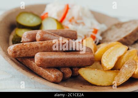 Saucisses savoureuses avec pommes de terre frites, concombre, chou et chou dans une assiette en papier sur une table en bois. Plats à emporter Banque D'Images