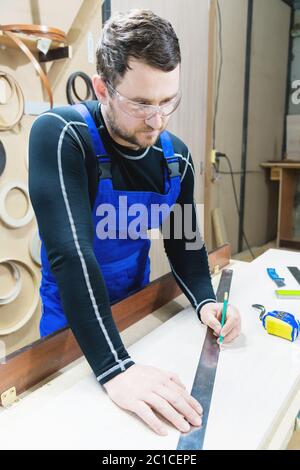 Un charpentier barbu fatigué sur une table avec un crayon dessine une enseigne sur le tableau. Sur le lieu de travail il y a un mas ordinaire Banque D'Images