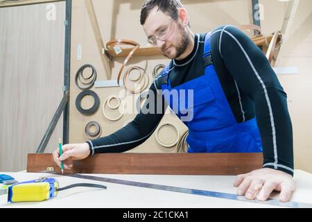 Un charpentier barbu fatigué sur une table avec un crayon dessine une enseigne sur le tableau. Sur le lieu de travail il y a un mas ordinaire Banque D'Images