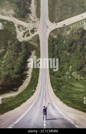 Un jeune homme en casquette avec une main levée avec une console d'un drone est debout sur une route de campagne asphaltée à la fin de W Banque D'Images