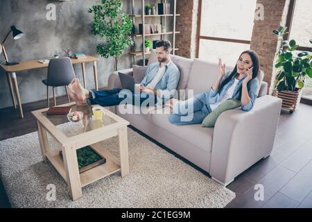 Photo pleine longueur de deux personnes homme travail à distance ordinateur portable SMS chat femme assis confort canapé appeler ami smartphone parler dire parler dedans Banque D'Images