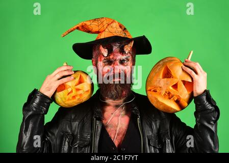 Démon avec des cornes et le visage strict tient Jack o lanternes. Homme portant un maquillage effrayant et un chapeau de sorcières tient des citrouilles sur fond vert. Halloween – concept de fête effrayante. Monstre avec décorations d'octobre Banque D'Images