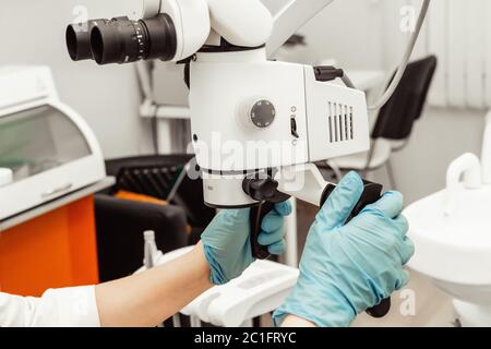 Jeune femme dentiste médecin regarde par un microscope professionnel dans une clinique dentaire. Un médecin dans un masque médical jetable et un capuchon. Avancé Banque D'Images