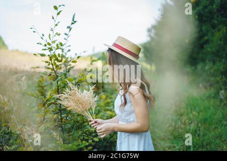 Belle fille dans un conseil et chapeau recueille des fleurs. La fille recueille un bouquet de fleurs séchées. Brunette fille parmi la grande herbe verte Banque D'Images