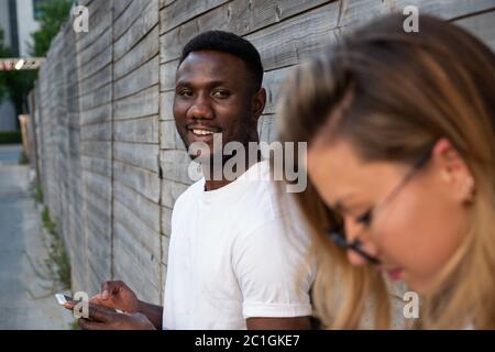 Jeune homme noir souriant devant un appareil photo près d'un mur à l'extérieur. Femme au premier plan. Se concentrer sur l'arrière-plan. Banque D'Images
