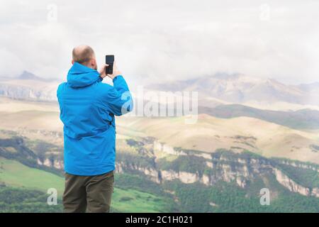 Un homme barbu dans une veste bleue à membrane se dresse dans la nature sur fond de montagnes et prend des photos sur le téléphone Banque D'Images