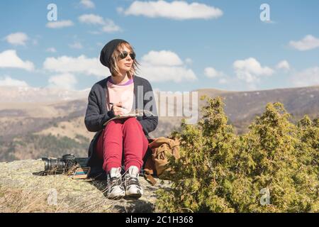 Une élégante petite fille peintre dans les montagnes, assise sur un rocher, s'inspire d'un bloc-notes au coucher du soleil Banque D'Images