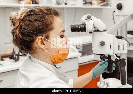 Jeune femme dentiste médecin regarde par un microscope professionnel dans une clinique dentaire. Un médecin dans un masque médical jetable et un capuchon. Avancé Banque D'Images