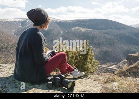 Portrait d'une fille de photographe de couturier hippster en chapeau et lunettes de soleil dessine un pastel dans son carnet assis sur un rocher en plein air Banque D'Images