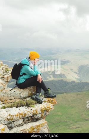 Jeune femme avec un sac à dos pensivement assis sur le bord d'un rocher et regardant le ciel avec des nuages Banque D'Images