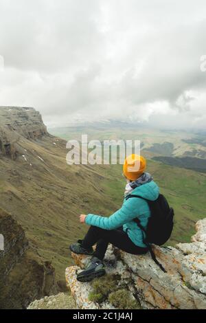 Jeune femme avec un sac à dos pensivement assis sur le bord d'un rocher et regardant le ciel avec des nuages Banque D'Images