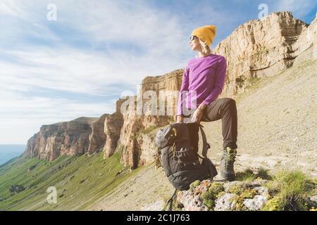 Une fille souriante voyageur dans un chapeau jaune et une paire de lunettes de soleil se tient au pied de roches épiques avec un sac à dos à côté et regarde un Banque D'Images