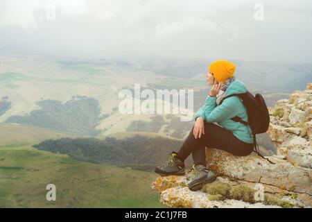 Jeune femme avec un sac à dos pensivement assis sur le bord d'un rocher et regardant le ciel avec des nuages Banque D'Images