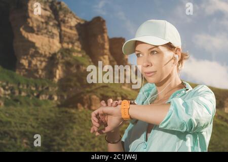 Portrait d'une jeune fille souriante en forme de fitness dans un casque et un casque vérifiant son horloge intelligente tout en étant assis à l'extérieur contre un dos Banque D'Images