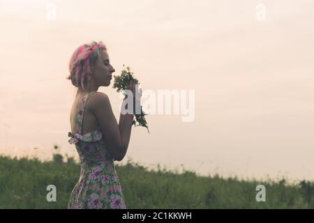 Jeune femme vêtue avec bouquet de fleurs entre les mains au coucher du soleil dans le champ. Image froide teintée Banque D'Images