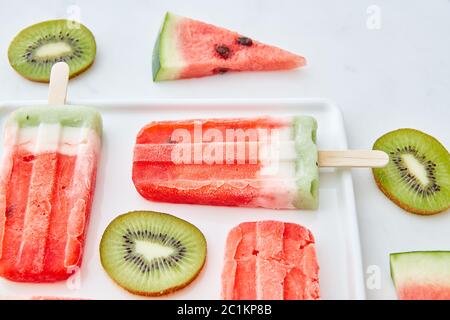 Crème glacée à la pastèque sur un bâton présenté avec des morceaux de kiwi et des morceaux juteux de pastèque dans une assiette sur un backgro en marbre gris Banque D'Images