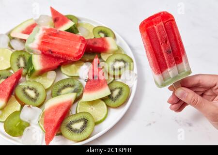 La main d'une femme tient une glace sur un bâton sous forme d'un morceau de pastèque sur un fond d'une assiette avec des morceaux de diff Banque D'Images