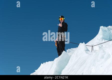 Un alpiniste se tient au bord d'un glacier avec une pelle à neige entre ses mains et montre le geste de Shak contre le ciel bleu Banque D'Images