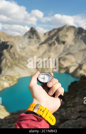 La main d'un homme tient un compas magnétique de poche pour la navigation sur fond de pente rocheuse et d'un lac de montagne. La conce Banque D'Images