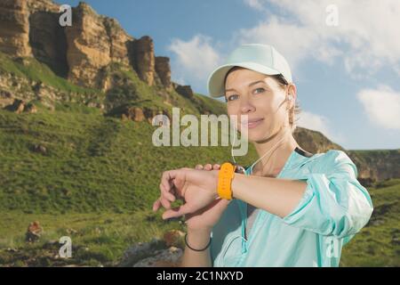 Portrait d'une jeune fille souriante en forme de fitness dans un casque et un casque vérifiant son horloge intelligente tout en étant assis à l'extérieur contre un dos Banque D'Images