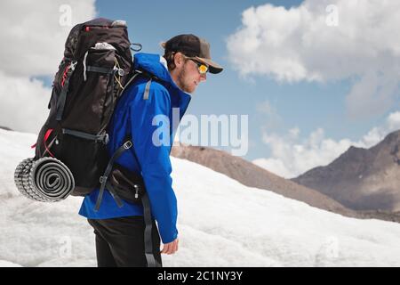 Voyageur dans une casquette et des lunettes de soleil avec un sac à dos sur ses épaules dans les montagnes enneigées sur le glacier contre le ciel et le clou Banque D'Images