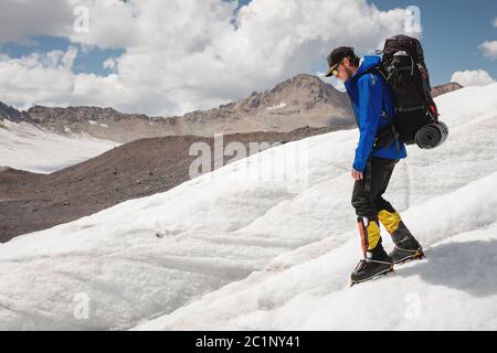 Voyageur dans une casquette et des lunettes de soleil avec un sac à dos sur ses épaules dans les montagnes enneigées sur le glacier contre le ciel et le clou Banque D'Images