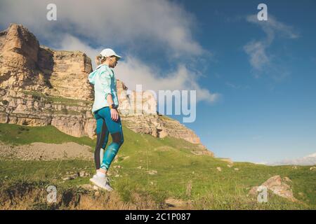 Une jeune fille sportive attirante dans un casque et un casque avant de courir dans un endroit pittoresque à côté des rochers au coucher du soleil. Entraînement Banque D'Images