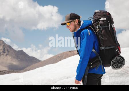 Voyageur dans une casquette et des lunettes de soleil avec un sac à dos sur ses épaules dans les montagnes enneigées sur le glacier contre le ciel et le clou Banque D'Images