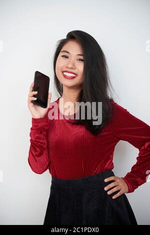 Portrait vertical moyen en studio d'une jeune femme asiatique attrayante tenant un smartphone moderne regardant l'appareil photo sourire, fond blanc Banque D'Images