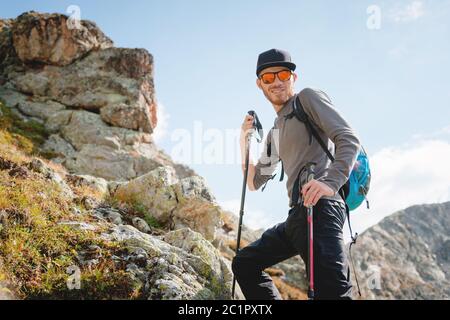 Porter d'un voyageur hippster élégant avec une barbe et un sac à dos en lunettes de soleil et une casquette avec des bâtons de randonnée se dresse sur un rock ag Banque D'Images