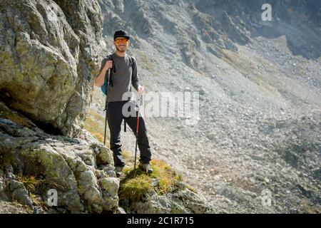 Porter d'un voyageur hippster élégant avec une barbe et un sac à dos en lunettes de soleil et une casquette avec des bâtons de randonnée se dresse sur un rock ag Banque D'Images