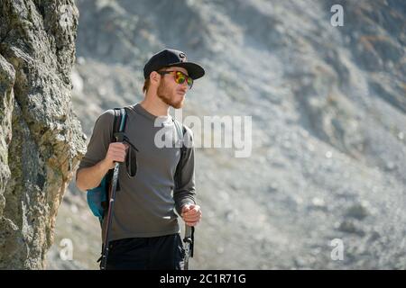 Porter d'un voyageur hippster élégant avec une barbe et un sac à dos en lunettes de soleil et une casquette avec des bâtons de randonnée se dresse sur un rock ag Banque D'Images