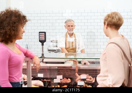 Vue arrière de deux femmes souriantes qui choisissent de la viande fraîche en magasin. Les femmes se tenant près du comptoir du magasin avec un grand assortiment de produits de viande crue, souriant maître d'hôtel senior montrant des saucisses et regardant l'appareil photo. Banque D'Images