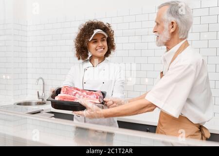 Foyer sélectif de jeune femme souriante avec cheveux bouclés recevant de la viande fraîche brute rouge dans un bol de boucher mâle senior. Fille gaie travaillant derrière le comptoir du magasin dans le supermarché. Banque D'Images